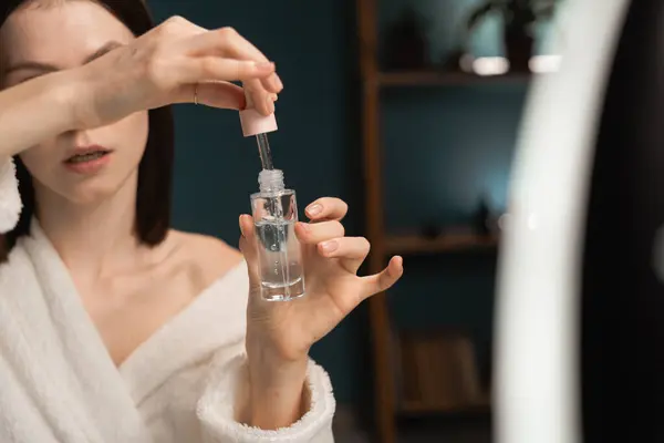 Skincare at home. Lovely beauty blogger in bathrobe present serum for skin rejuvenation in home interior. Influencer recording video review blog using ring lamp and smartphone on tripod. Copy space