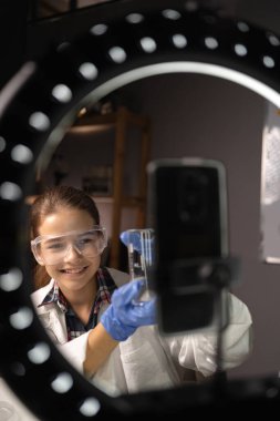 Küçük bayan blogcu, takipçilerine laboratuarında kimya deneyi yaparken sıvılarla test tüpünü gösteriyor. Günlük konsepti