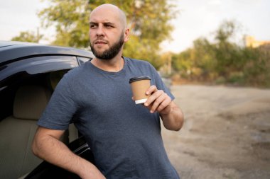 Erkek sürücü arabasının yanında dikilirken paket kahve içiyor. Sürücü ve tatil kavramı