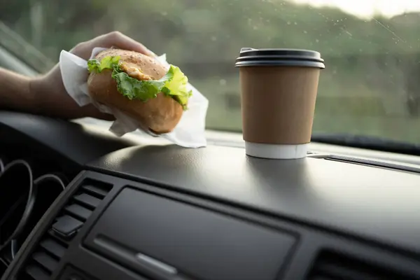 Male hands holding hamburger and cup of coffee in car, dangerous and risk an accident. Copy space