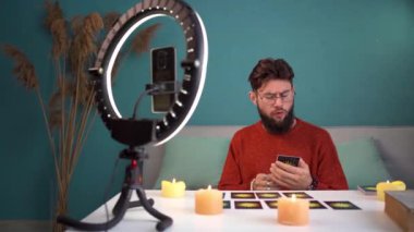Profesyonel internet flaması Tarot kartları kullanarak fal bakar. Video yayını için akıllı telefonu ve halka lambasını kullanarak canlı yayın. Kameranın karşısına geçmiş ve şaşırmış bir şekilde bir kaç kart çekmiş.