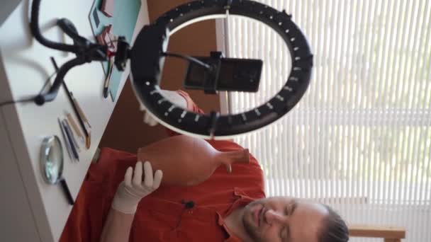 一位留着胡子的高加索男性考古学家博主正在他的工作室里用智能手机和戒指灯录制这个节目 显示一个旧花瓶正在拆卸 垂直录像 — 图库视频影像