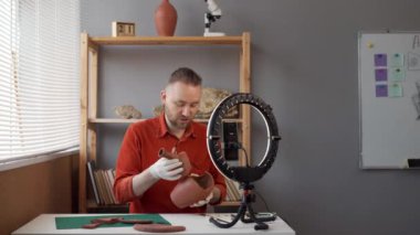 Sakallı beyaz erkek arkeolog blogcu atölyesinde akıllı telefon ve yüzük lambası kullanarak bir video kaydediyor. Eski bir vazonun parçalara ayrıldığını ve detaylara odaklandığını gösteriyor. Boşluğu kopyala.