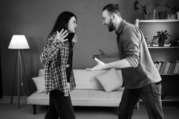 Black and white photo of angry couple fighting and shouting at each other in the living room at home. Copy space