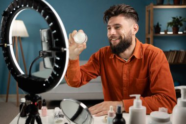 Video kaydederken kozmetik ürünlerini gösteren ve akıllı telefon kamerasında güzellik blogu için tavsiyelerde bulunan ve halka lamba kullanan bir erkek vlogger. Boşluğu kopyala