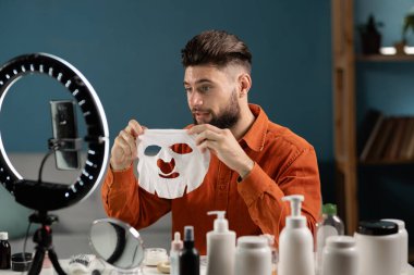 Cilt bakımının reklamını yapan yakışıklı adam kameraya kumaş maskesi gösteriyor. Güzellik günlüğü kavramı