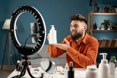 Tripodun üzerinde akıllı telefonun önünde oturan, evden yayın yapan, kozmetik ürünlerini tanıtan bir erkek güzellik uzmanı. Boşluğu kopyala
