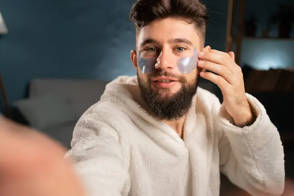 Man after a shower wears cosmetic patches under the eyes. Making selfie on cellphone. Cosmetic procedures at home. Getting ready for a sleep. Copy space