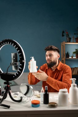 Erkek blogcu etkileyicisi video iletişimini çevrimiçi kaydediyor, krem tutuyor ve kozmetik ürünler hakkında konuşuyor. Boşluğu kopyala