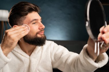 Çekici bir adam yüzünü temizliyor, pamuk pedlerle ve temizlik ürünleriyle aynaya bakıyor. Evin içinde yüz toneri ve pamuk pedi kullanan bir adam. Boşluğu kopyala