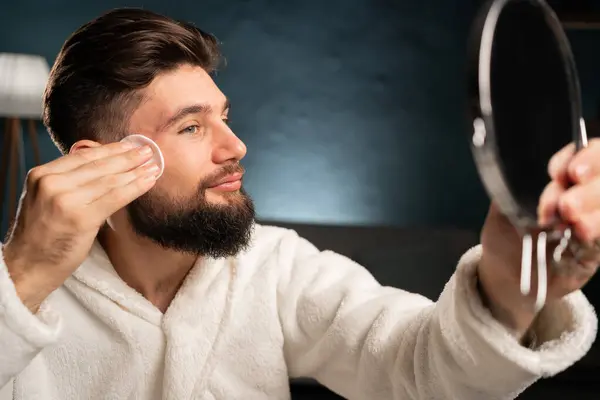 Attractive man cleaning his face, using cotton pads and cleansing product, looking at mirror. Guy using face toner and cotton pad in home interior. Copy space