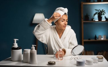 Evde güzellik prosedürü. Gözünün yanında kırışıklık önleyici serumla pipette tutan bornoz giymiş aynaya bakan, akşamları yatak odasında oturan İspanyol kadın.