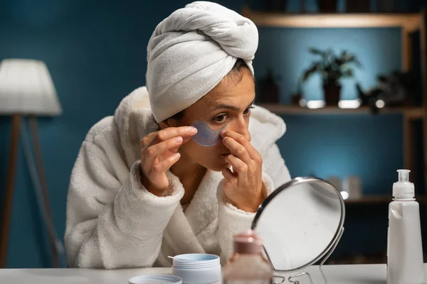 Hygiene, beautician, makeup and face care concept. Patches for eyes. Hispanic woman with towel on her head glues transparent patches to skin under her eyes. Copy space