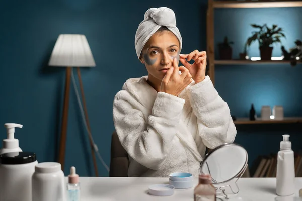 Millennial woman applying under eye patch on face and looking at camera. Beautiful woman wear bathrobe and wrapped bath towel on head. Face skin care. Copy space