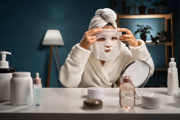 Beautiful lady applying cotton face mask sitting at table with various homemade cosmetics. Woman using cosmetic for facial treatment. Copy space