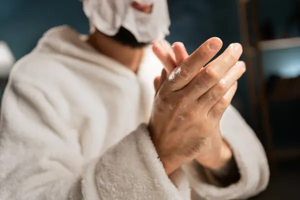 A young man taking care of the skin of his hands in the evening in the bedroom applies moisturizing cream to his hands for delicate skin. Close-up