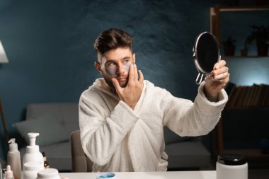 Genç adam duş aldıktan sonra oturma odasında anti-yaş rutininin tadını çıkarıyor. Bornoz giymiş bir adam göz altına hidrojel bantlar yapıştırıyor, aynaya bakıyor. Boşluğu kopyala