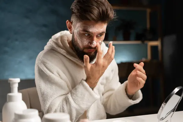 Man applying moisturizer to his face while sitting in the living room in the evening after a shower looking in the mirror while taking care of his skin. Copy space