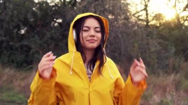 Sarı yağmurluk giyen, tarlada dans eden ve kulaklıkla müzik dinleyen güzel bir kadın. Yaz yağmurunun ve dansın tadını çıkaran bir kız. Boşluğu kopyala