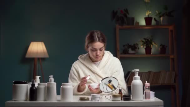 年轻的女人穿着浴衣 在脸上涂上乳霜 晚上在家里照镜子 美容美发 皮肤护理和人的观念 复制空间 — 图库视频影像