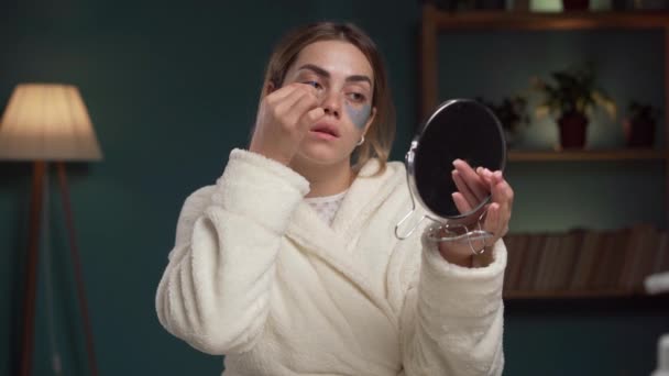 穿着浴衣的年轻女子用眼部胶原补丁贴着镜子 在眼罩下使用抗疲劳药的女孩 复制空间 — 图库视频影像