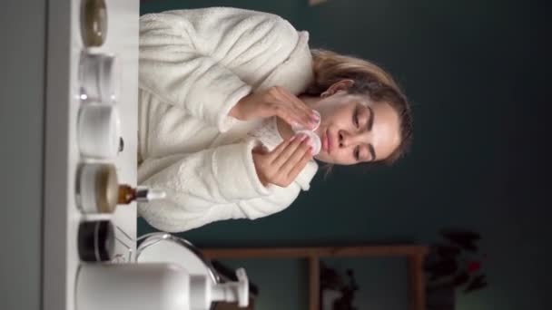 女人在家里用棉垫和胶水擦拭脸 用棉垫和镜子坐在一起 面部皮肤护理常规的概念 垂直录像 — 图库视频影像
