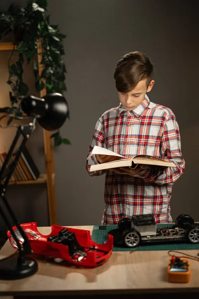 Focused boy reading science book and using soldering iron for fixing remote controlled toy car at home. Copy space