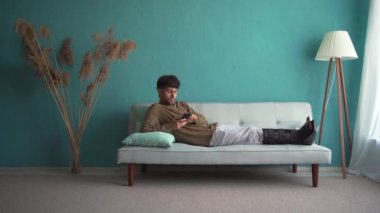 Kanepeye uzanmış akıllı telefonlu, yürüyen telli Arap bir genç. Ayak bileği ortozu. Boşluğu kopyala