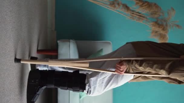 足首のブレースブーツを歩き 怪我の後 自宅でクラッチを歩いている若者 バーティカルビデオ — ストック動画