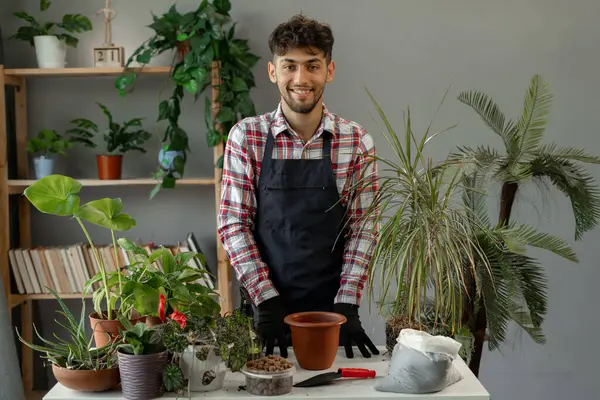 A young Arab man replanting house plants into new pots looks at the camera and smiles. Home hobby. Copy space