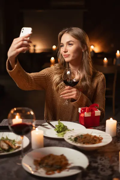 Beautiful young woman having a romantic candlelight dinner at home, making selfie on smartphone, drinking wine. Copy space