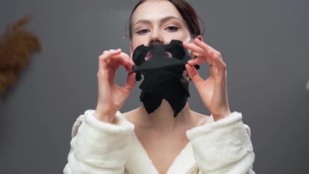 魅力的な若い女性は カメラを見ている彼女の顔からシートマスクを浄化しています フェイススキンケアコンセプト — ストック動画