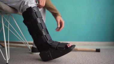 Evdeki sandalyede ayak bileği ortozu olan bir adam. Yürüteç botlu yaralı adam. Boşluğu kopyala