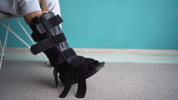 男人穿上脚踝支架 整形外科脚踝支撑鞋 受伤的家伙在椅子上与斯普林特 步行靴 脚踝矫形术 复制空间 — 图库视频影像