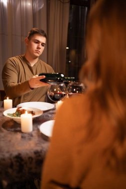 Aşık genç bir çift şarap içiyor, adam bardaklara şarap dolduruyor sevgililer gününü evde yemek yiyor, mumlarla romantik bir akşam yemeği yiyor. Boşluğu kopyala