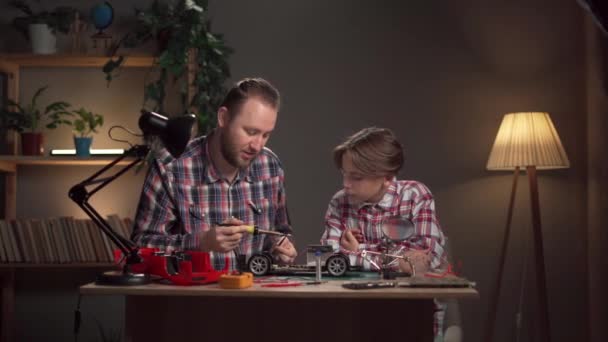 父亲和儿子坐在桌旁 用焊锡熨斗修理一辆受控玩具汽车 复制空间 — 图库视频影像
