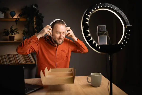 Handsome man recording video on phone camera while unpacking box with new wireless headphone, testing at home. Male influencer sharing with subscribers feedback about new headset. Copy space