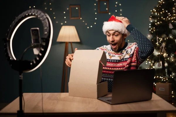 快乐的穆斯林男性博主 戴着圣诞老人帽打开礼物盒 进行现场直播 用智能手机相机为社交网络创建内容的阿拉伯人 复制空间 图库图片