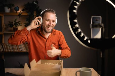 Yakışıklı beyaz adam, evinde yeni kablosuz kulaklıklarla kutusunu açarken telefon kamerasıyla video kaydediyor. Etkileyici, yeni teknoloji ürünü incelemesi. Boşluğu kopyala