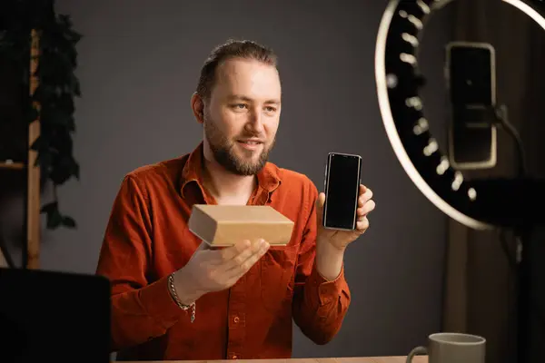 Concept of people, technology and blogging. Handsome man filming video on phone camera while opening parcel box with new smartphone at home. Copy space