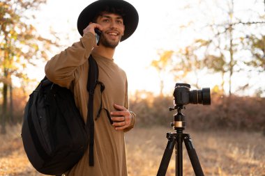 Sırt çantası, kamera ve tripodla çevrili Arap genç fotoğrafçı cep telefonundan arıyor. Gezgin turist profesyonel videograf macera tatili çekimleri slr kamera.