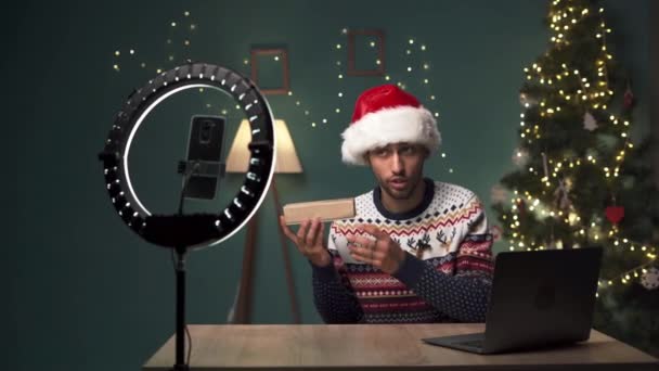 阿拉伯男性博主打开新的智能手机 并在相机上录制视频 年轻人坐在圣诞节的房间里 戴着圣诞帽与订户分享他对现代小玩意的反馈 — 图库视频影像