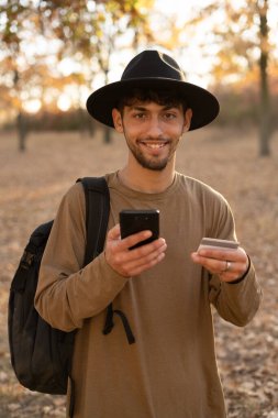 Telefon ve kredi kartı kullanan genç bir Arap. Ormanda dikilip kameraya gülümsüyorsun. Portre