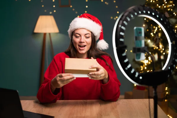 Video review. Young Caucasian woman showing cardboard box in front of smartphone camera, smiling recording content for her fashion blog, unpacking box while sitting on sofa at home wearing santa hat.