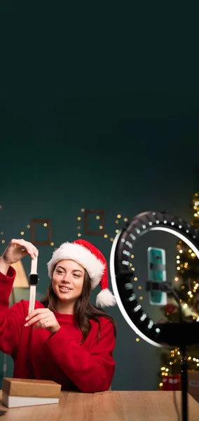 Video review. Young Caucasian woman demonstrating smart watch in front of smartphone camera, recording content for her fashion blog, unpacking box, sitting at home wearing santa hat. copy space.