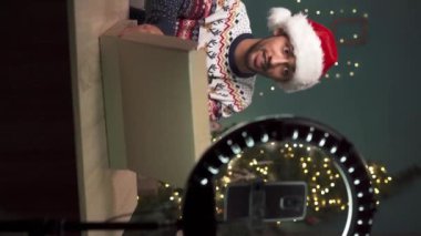 Arap erkek blogcu Noel Baba şapkalı ve yeni dizüstü bilgisayarlı paketini açarken Noel arifesinde evde teknoloji blogu için video içeriği kaydediyor ve tripoda akıllı telefon kullanıyor. Dikey video