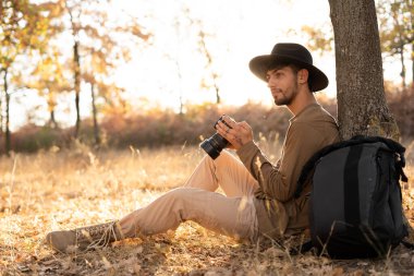 Sırt çantalı ve kameralı bir adam ağacın altında oturuyor ve sarı parkın keyfini çıkarıyor. Turist sonbahar ormanında dinleniyor ve dinleniyor. Boşluğu kopyala