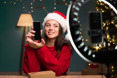 Boks videosu açılıyor. Evde Noel Baba şapkalı beyaz bir kadın abonelerine canlı yayın sırasında yeni bir akıllı telefon gösteriyor. Etkilendirici reklam konsepti. Pankart. Boşluğu kopyala.