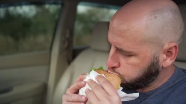 Beyaz adam arabada lezzetli sıcak burger yiyor, tehlikeli ve kaza riski var. Yakın plan.