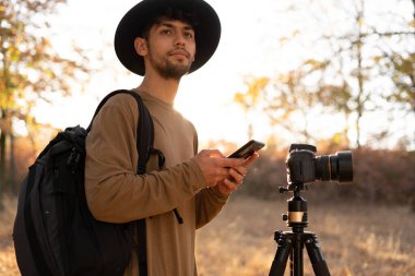 Doğa fotoğrafçısı güneş doğarken kamerayla fotoğraf çeker, akıllı telefon kullanır ve başka yere bakar. Boşluğu kopyala